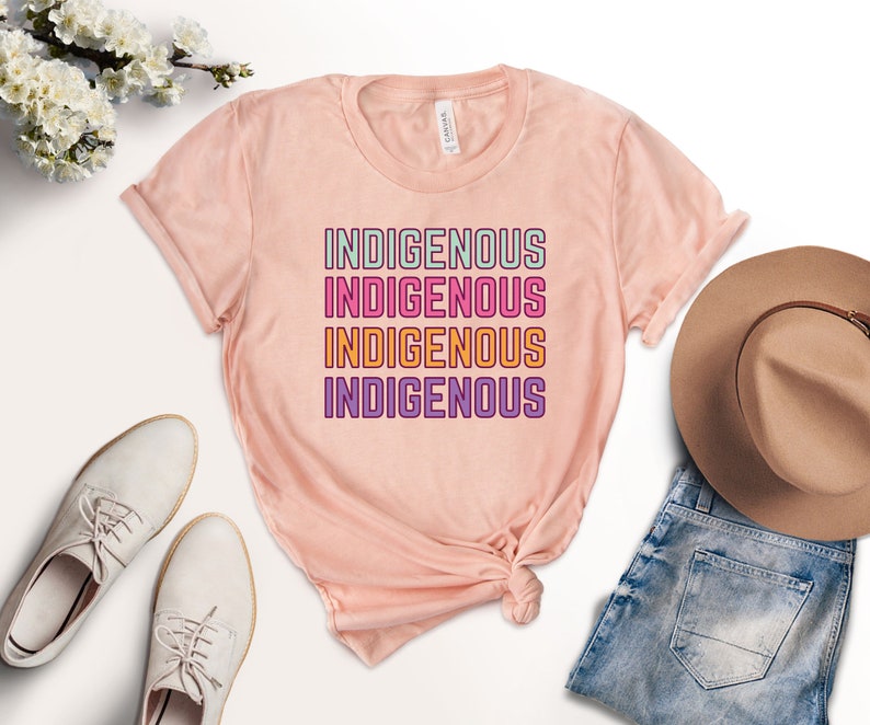 May include: A pink t-shirt with the word "INDIGENOUS" repeated four times in a stacked design. Each word is in a different color: green, pink, orange, and purple.