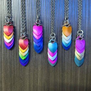 May include: Six necklaces featuring pendants made of layered, scale-shaped metal plates in various vibrant colors including red, orange, yellow, green, blue, purple, and pink, all suspended from silver-toned metal chains with circular jump rings.