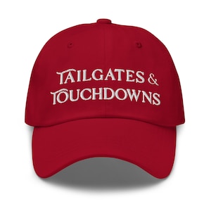 Könnte beinhalten: Rote Baseballkappe mit dem weißen, gestickten Text "TAILGATES & TOUCHDOWNS". Die Kappe hat einen gebogenen Schirm und ein klassisches Sechs-Panel-Design. Ein Accessoire für Sportfans.