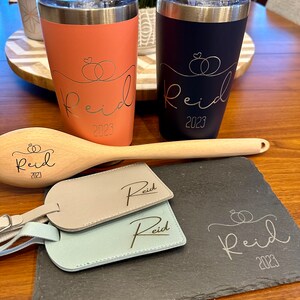 Puede incluir: Dos vasos de acero inoxidable con un exterior negro y coral. Ambos vasos tienen el nombre "Reid" y el a&ntilde;o "2023" grabados. Los vasos est&aacute;n sobre una superficie de madera con una cuchara de madera y dos etiquetas de equipaje de cuero con el nombre "Reid" grabado. Una bandeja de servir de pizarra negra con el nombre "Reid" y el a&ntilde;o "2023" grabados est&aacute; en primer plano.