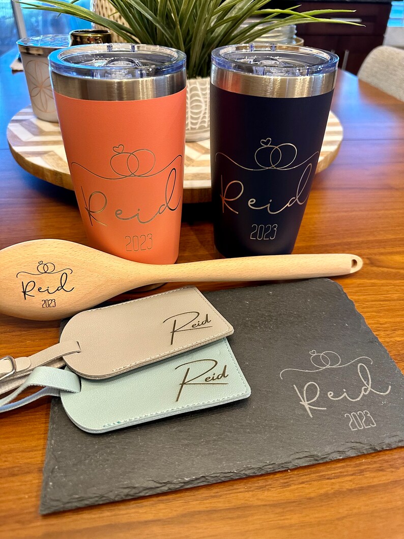 Puede incluir: Dos vasos de acero inoxidable con un exterior negro y coral. Ambos vasos tienen el nombre "Reid" y el a&ntilde;o "2023" grabados. Los vasos est&aacute;n sobre una superficie de madera con una cuchara de madera y dos etiquetas de equipaje de cuero con el nombre "Reid" grabado. Una bandeja de servir de pizarra negra con el nombre "Reid" y el a&ntilde;o "2023" grabados est&aacute; en primer plano.
