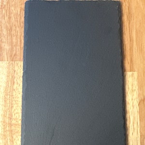 Puede incluir: Una tabla de servir de pizarra negra con una superficie rugosa y texturizada. La tabla es rectangular y tiene bordes ligeramente redondeados.