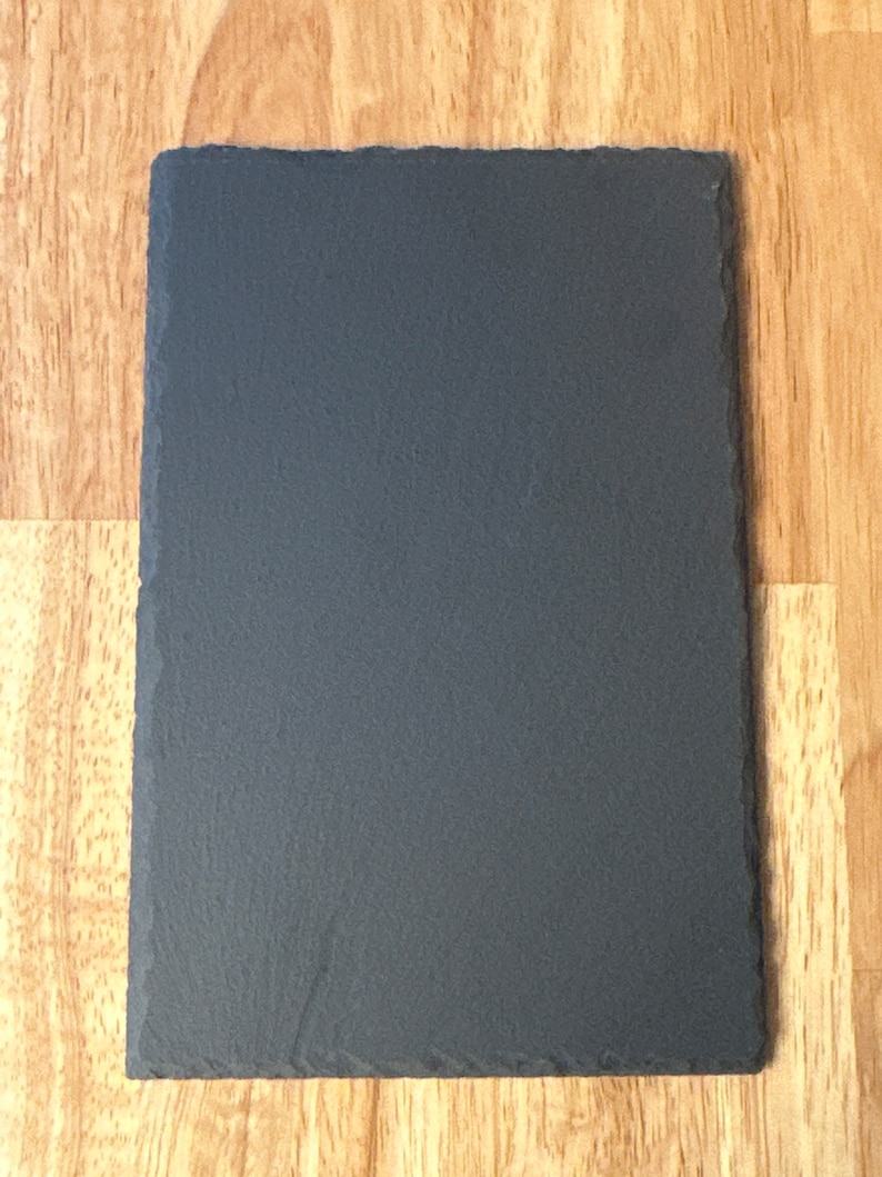 Puede incluir: Una tabla de servir de pizarra negra con una superficie rugosa y texturizada. La tabla es rectangular y tiene bordes ligeramente redondeados.