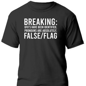 May include: Black t-shirt with white text that reads "BREAKING: UFO'S HAVE BEEN IDENTIFIED, PRONOUNS ARE ABSOLUTELY, FALSE/FLAG"