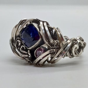 May include: A silver ring with a blue gemstone and two smaller pink gemstones. The ring is intricately designed with wire wrapped around the stones.