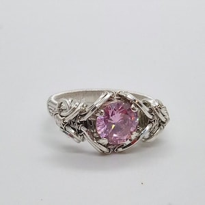 May include: A silver-coloured wire-wrapped ring with a large, round, light pink gemstone. The ring's band and setting are intricately woven with wire, creating a detailed and textured design. The gemstone is the focal point.