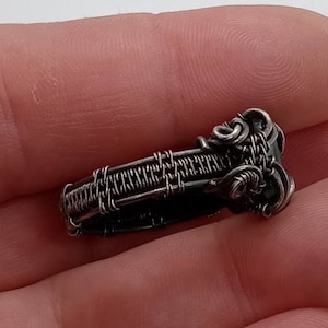 May include: A handcrafted silver wire-wrapped pendant. The pendant features intricate wire weaving and decorative swirls. The design is a small, rectangular shape with a dark stone or bead accent. The pendant is held in a hand against a white background.