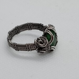 May include: A handcrafted wire-wrapped ring with a dark silver finish. The ring features a faceted, oval-shaped green gemstone. The wirework creates a textured band and a decorative setting for the stone, showcasing intricate details.