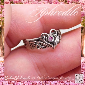 Może przedstawiać: Srebrny pierścionek owinięty drutem z wzorem różowego serca i małym różowym kamieniem szlachetnym w środku. Pierścionek jest na palcu, a słowo "Aphrodite" jest napisane kursywą nad pierścionkiem.