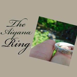 Könnte beinhalten: Ein silberner und goldener drahtgewickelter Ring, bekannt als "Aiyana Ring", wird in einer Hand vor einem verschwommenen Hintergrund aus grünem Laub gehalten.