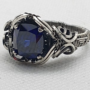 May include: A silver ring with a large, square-cut, deep blue gemstone. The gemstone is set in a detailed, wire-wrapped design with ornate swirls and patterns. The ring's band is intricately woven with wire.
