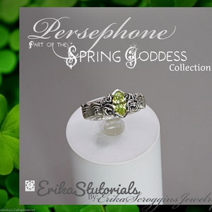 May include: A silver ring with an oval, light green gemstone at the center. The ring is part of the Spring Goddess Collection, with the name "Persephone" above it. The ring has intricate floral and scrollwork designs on the band.