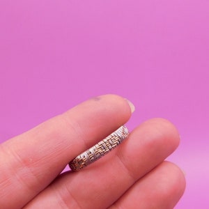 May include: A silver and gold ring with a woven design. The ring is held between two fingers against a pink background. The ring's intricate pattern features alternating silver and gold bands.