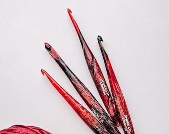 Puede incluir: Cuatro agujas de crochet rojas y negras con el texto "OPHIRE CO. 4.5mm(G)" y "OPHIRE CO. 6.5mm(K)" en las agujas. Dos ovillos de lana, uno rojo y uno negro.