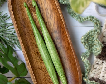 Puede incluir: Dos ganchillos de color verde claro con un acabado liso y pulido y una sutil textura de veta de madera, que descansan dentro de un cuenco de madera alargado de color marrón oscuro con un patrón de veta natural visible.