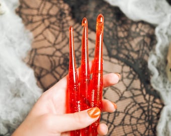 Pode incluir: Três agulhas de crochê vermelhas translúcidas com partículas de glitter incorporadas. Cada agulha apresenta uma haste cônica e uma ponta afiada. O texto OPHIRE CO e 6.0mm está impresso em branco na superfície das agulhas, que são seguradas juntas.