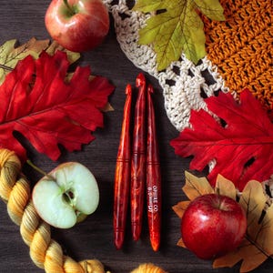 May include: Three red marbled crochet hooks with the text OPHIRE CO. 6.0mm (J) printed on one hook, surrounded by two whole red apples, one halved apple, a yellow yarn skein, a cream crocheted fabric piece, and several autumn-colored leaves.