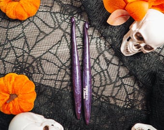 May include: Two purple crochet hooks with a marbled finish. The right hook features white text that reads OPHIRE CO. 7.0mm. The hooks have a tapered design with a pointed tip and a smooth, rounded handle, showcasing a consistent deep violet color.
