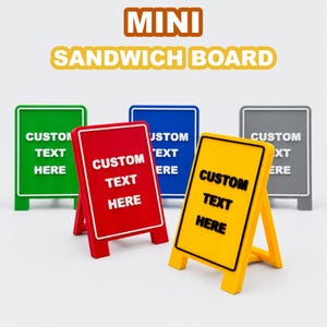 Peut inclure: Un ensemble de cinq mini panneaux publicitaires de type chevalet en vert, rouge, bleu, jaune et gris. Chaque panneau présente une bordure en relief blanche ou noire et le texte CUSTOM TEXT HERE en lettres en relief assorties.