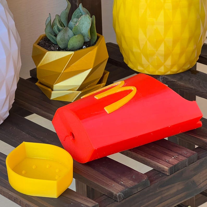 Może przedstawiać: Czerwona i ż&oacute;łta dekoracyjna donica w kształcie pojemnika na frytki McDonald's. Donica ma logo McDonald's z boku. Inne donice na zdjęciu to złota geometryczna donica z sukulentem i ż&oacute;łta donica w kształcie ananasa.