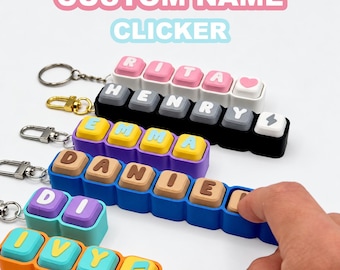 May include: A collection of custom name clicker keychains featuring rows of square, colorful buttons with letters and symbols. Each keychain has a metal ring attachment. The buttons display names like RITA, HENRY, EMMA, DANIEL, DI, and IVY in various colors.