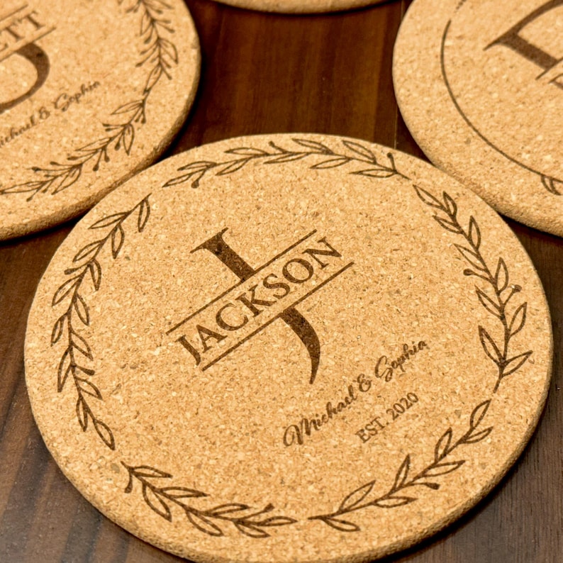 May include: Cork coaster with a wreath design and the name "JACKSON" in a banner with the initial "J" in a larger font. The text "Michael & Sophia EST. 2020" is also engraved on the coaster.