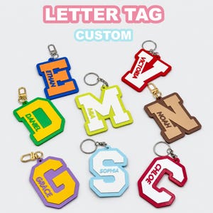 May include: A collection of custom letter keychains featuring various colors and names including Ethan, Daniel, Mia, Victoria, Noah, Grace, Sophia, and Chloe. Each tag has a metal keyring and displays a large varsity-style letter with a name.