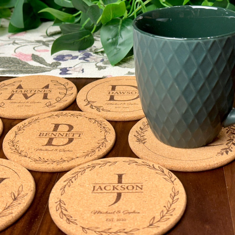 May include: Set of six cork coasters with personalized names and initials. Each coaster features a wreath design with a leafy border. The coasters are displayed on a wooden surface with a green mug.