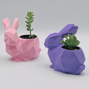 May include: Two geometric bunny shaped planters, one pink and one purple, with small green plants inside.