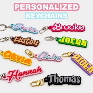 May include: A collection of personalized keychains featuring names such as Sophia, Brooke, Easton, Jacob, Olivia, Claire, Aiden, Hannah, and Thomas in various colors and fonts, each attached to a metal keyring or a gold-toned lobster clasp.