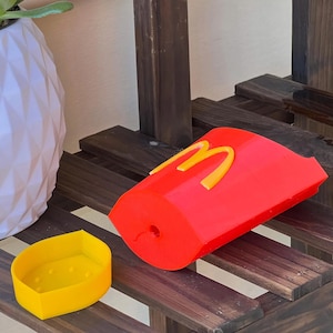 Może przedstawiać: Czerwony pojemnik drukowany w 3D z ż&oacute;łtym logo "M", przypominający pudełko na frytki z fast foodu, oraz osobny ż&oacute;łty pojemnik. Przedmioty znajdują się na drewnianej powierzchni.