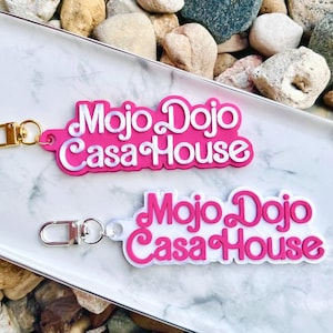 Puede incluir: Dos llaveros rosa y blanco con el texto "Mojo Dojo Casa House" en letras blancas. Los llaveros están hechos de un material impreso en 3D.