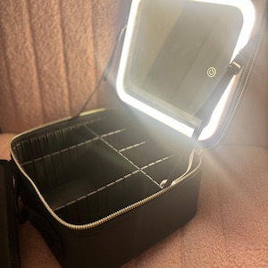 May include: Black makeup case with a built-in mirror and a ring light. The case has multiple compartments for storing makeup and other beauty supplies.