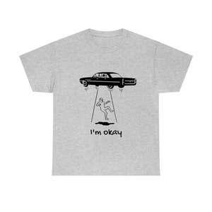 May include: A gray t-shirt with a black illustration of a car flying in the air with beams of light coming from the bottom of the car. A person is standing on the ground below the car with the text "I'm okay" printed below the person.