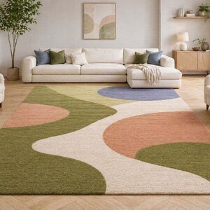 K&ouml;nnte beinhalten: Ein gro&szlig;er Teppich mit abstraktem Design in Gr&uuml;n-, Pfirsich-, Blau- und Cremet&ouml;nen. Der Teppich befindet sich in einem Wohnzimmer mit einem wei&szlig;en Sofa und Sesseln. Die Abmessungen des Teppichs sind nicht sichtbar.