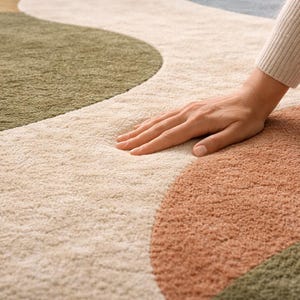 K&ouml;nnte beinhalten: Ein weicher Teppich mit abstraktem Muster in Beige, Olivgr&uuml;n und Terrakotta. Das Design zeigt gro&szlig;e, organische Formen. Eine Hand liegt auf dem Teppich und unterstreicht seine weiche Textur und einladende Haptik.