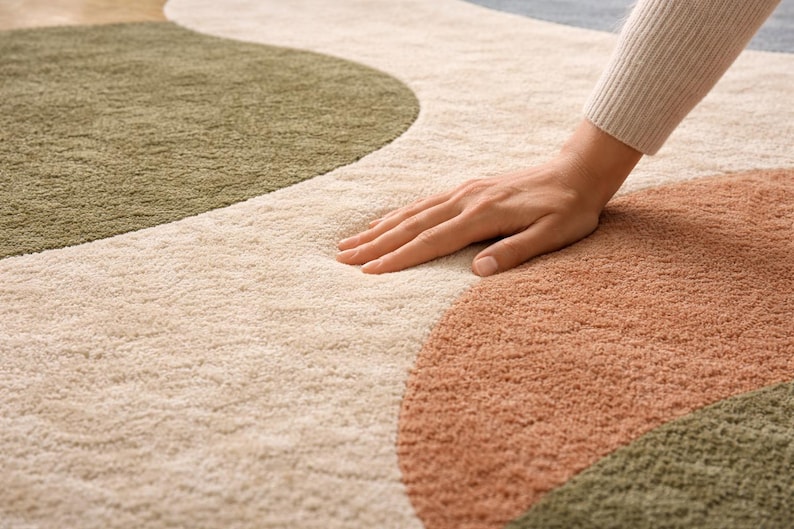 K&ouml;nnte beinhalten: Ein weicher Teppich mit abstraktem Muster in Beige, Olivgr&uuml;n und Terrakotta. Das Design zeigt gro&szlig;e, organische Formen. Eine Hand liegt auf dem Teppich und unterstreicht seine weiche Textur und einladende Haptik.