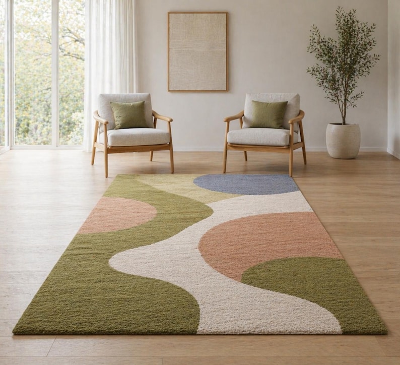 K&ouml;nnte beinhalten: Rechteckiger Teppich mit abstraktem Design in Gr&uuml;n, Rosa, Blau und Creme. Der Teppich hat ein wellenf&ouml;rmiges Muster und liegt auf einem Holzboden. Im Hintergrund befinden sich zwei Sessel mit olivgr&uuml;nen Kissen.