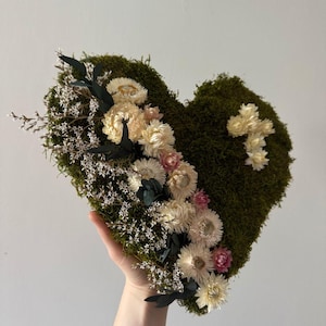 Könnte beinhalten: Eine herzförmige Wanddekoration aus dunkelgrünem Moos mit einer diagonalen Anordnung aus cremefarbenen Trockenblumen, kleinen weißen sternförmigen Blüten und dunkelgrünen Blättern sowie einem kleinen Kreuz aus cremefarbenen Blüten rechts.