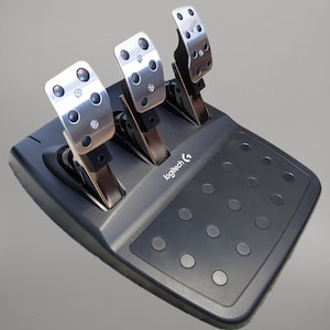 Può includere: Un set di pedali per giochi di corse nero e argento. Il set presenta tre pedali con facce in metallo argentato e dettagli in gomma nera. La base è nera con un'area poggiapiedi strutturata e il logo Logitech.