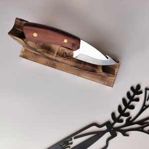 Op de afbeelding: Een houten messenhouder met een mes met een bruin houten handvat en een zilveren blad. Het mes zit in de houder. Er is een zwart metalen bloemmotief met paarse bloemen op het witte oppervlak.