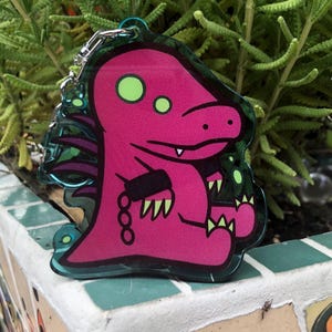 May include: Pink and green cartoon monster keychain with a chain and a black band on its arm. The monster has a toothy grin and is sitting with its legs crossed.