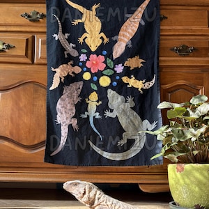May include: Black decorative wall hanging featuring a colorful illustration of various lizards and flowers. The lizards are in shades of yellow, brown, and gray. The flowers are pink, purple, and yellow. The text "DANIEMIZU" is printed on the wall hanging.