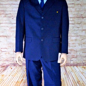 Puede incluir: Un traje azul marino exhibido en un maniquí. El traje incluye una chaqueta de tres botones, pantalones a juego, una camisa azul claro y una corbata azul marino. Un pequeño emblema amarillo está en la chaqueta.