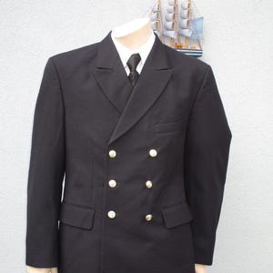 Könnte beinhalten: Ein schwarzer zweireihiger Blazer mit goldenen Knöpfen. Der Blazer hat einen hohen Kragen und einen einzigen Knopfverschluss oben.