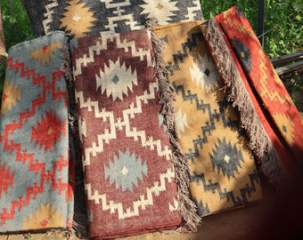 May include: A collection of colorful jute rugs with geometric patterns. The rugs feature designs in shades of gray, red, gold, and brown, with fringed edges. The rugs are displayed outdoors, leaning against a metal railing.