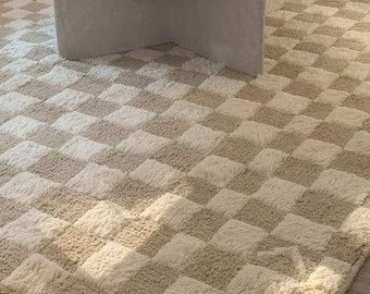 May include: A beige and cream checkered area rug in a living room setting. The rug is rectangular and covers a large portion of the floor. A round, light-colored coffee table and a pink sofa are in the background.