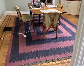 May include: A dining room scene featuring a wooden table with a laptop and placemats, surrounded by four chairs. A large rug with a pink and navy blue geometric pattern covers the floor. The room has a neutral color palette.