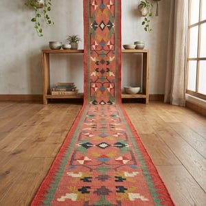 Könnte beinhalten: Ein langer, schmaler Läufer mit korallenroter Grundfarbe und einem sich wiederholenden geometrischen Muster in Grün-, Blau-, Gelb- und Weißtönen. Der Teppich hat einen Fransenabschluss und ein kleines rechteckiges Etikett mit der Aufschrift Handmade Kilims.