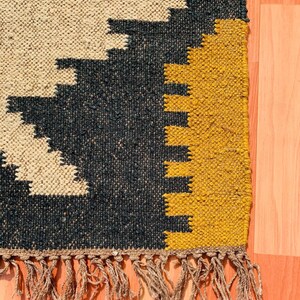 May include: Close-up of a woven rug with a geometric pattern. The rug features a beige background with black and yellow geometric shapes. The rug has a fringe along the bottom edge.